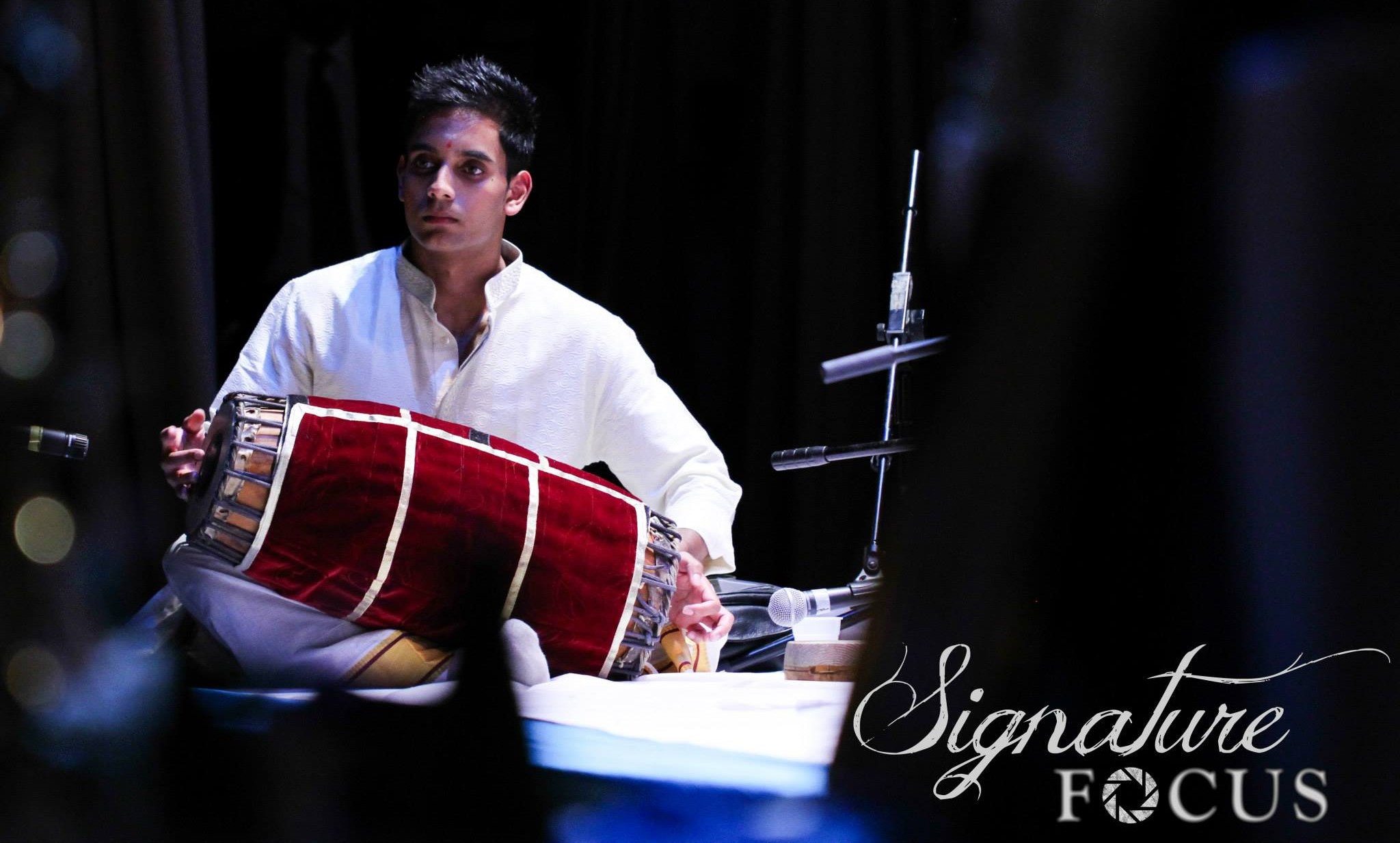 Abhiram Sahathevan, dressed in white, sits crosslegged on-stage, with a mridangam drum across his lap. The drum is wrapped in red velvet.