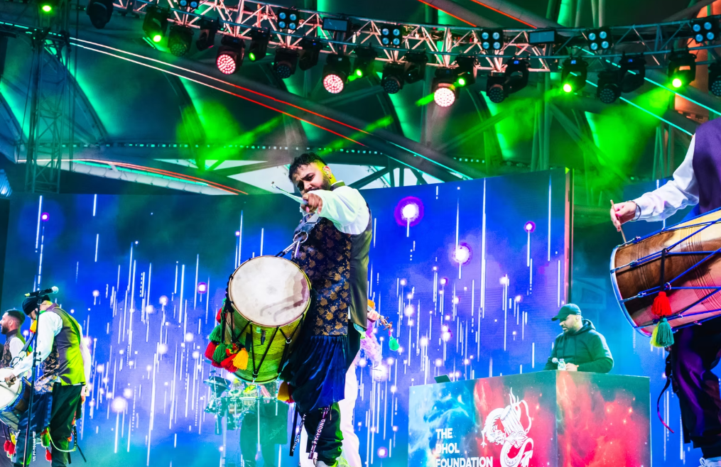 Three dhol players on a stage, shot from the pit - with colourful projected backdrop. the centre player points directly at the camera.